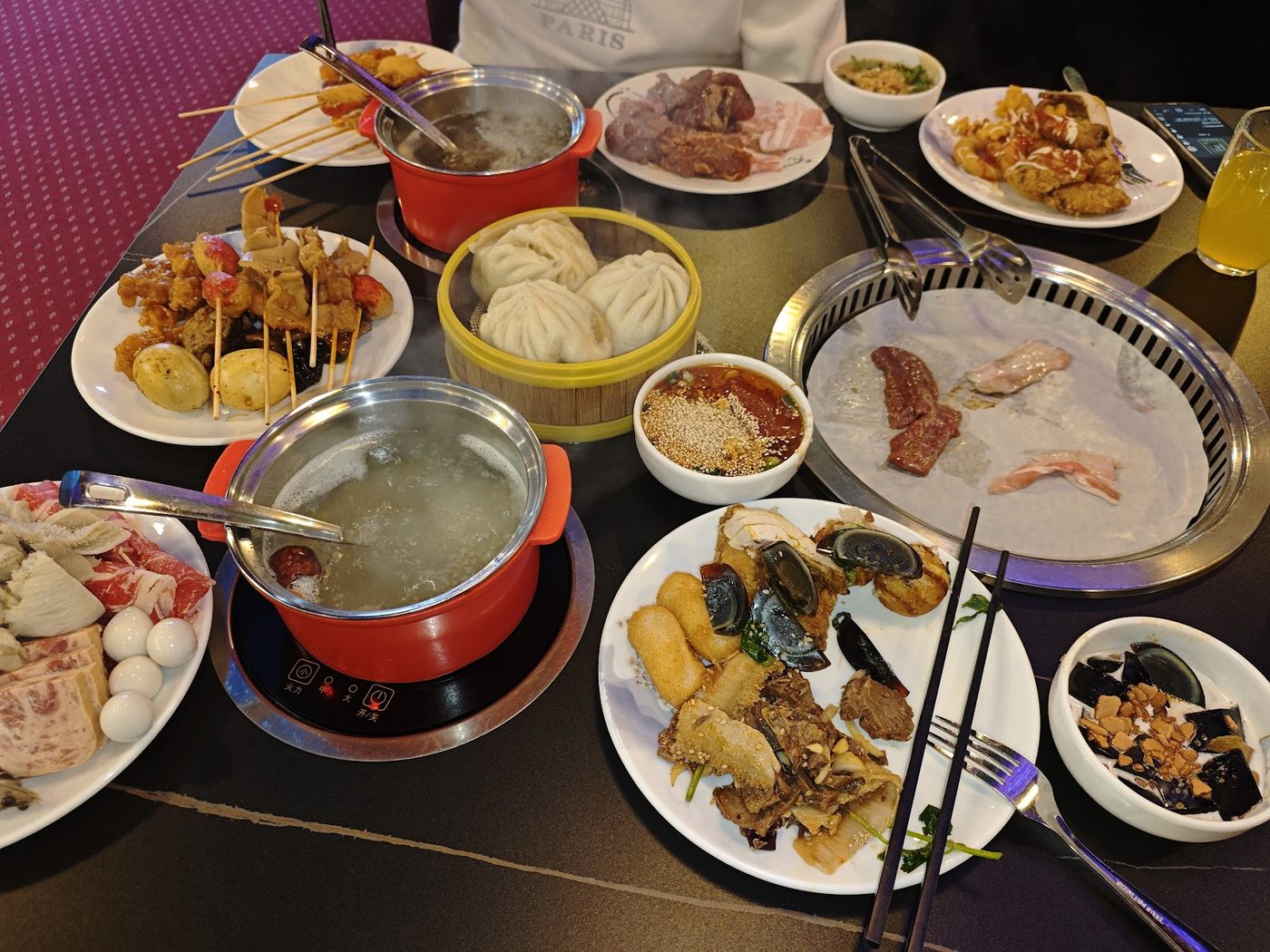 A table set with two pots of broth, a grill, a bamboo steamer of buns and skewers